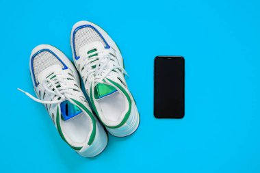 Mavi arka planda akıllı telefonlu parlak çizgilerle dekore edilmiş yeni beyaz ayakkabılar, üst manzara, kopyalama alanı. Lüks yaşam tarzı için modern aksesuarlar. Reklam afişi için stüdyo çekimi.