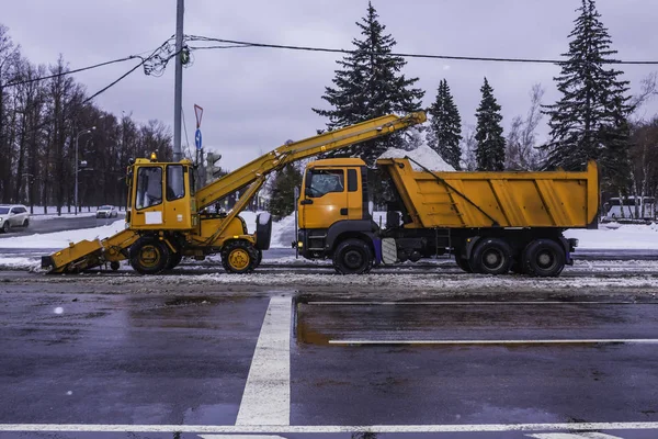 Machine shovels snow from the street and loading snow into a truck . Orange machines, snow cleaners , near traffic lights . Moscow , Russia .