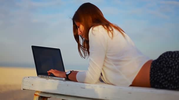 Женщина лежит на скамейке у моря и печатает на ноутбуке — стоковое видео