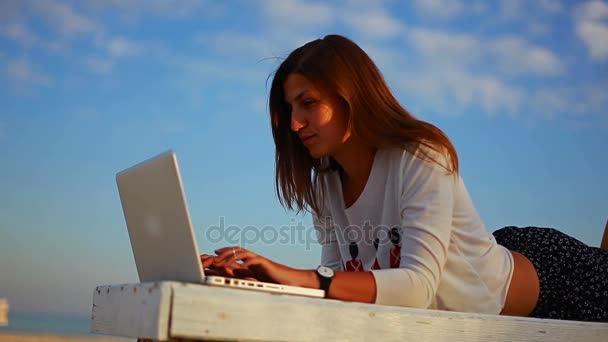 Молодая женщина работает онлайн, лежа на лавочке в выходные у моря — стоковое видео