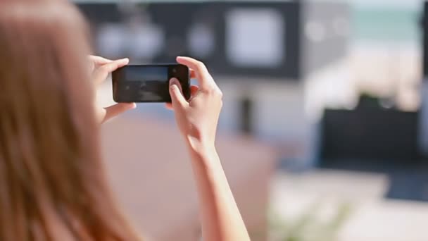 Девушка фотографирует современные пляжные домики по телефону — стоковое видео