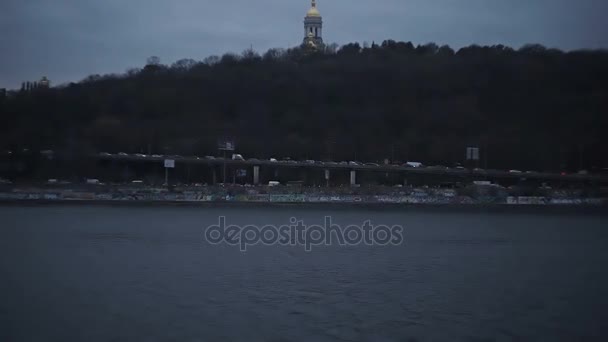 Drukke verkeer langs de waterkant in de avond, weergave van orthodoxe kerk van rivier — Stockvideo