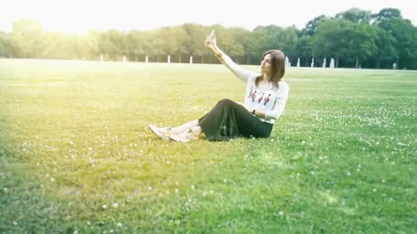 Çimenlerin üzerine oturmuş ve selfie smartphone yapma güzel kız — Stok video