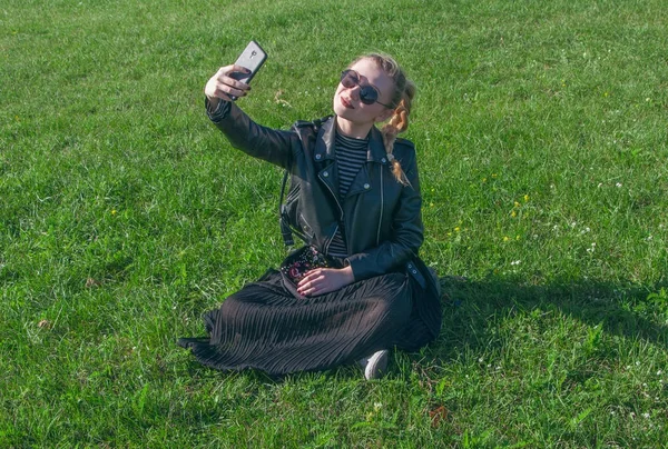 Hermosa chica rubia sentada en un césped verde y hace selfie — Foto de Stock