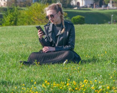 Güzel sarışın kız bir yeşil çimenlerin üzerinde oturur ve bir akıllı telefon görünüyor