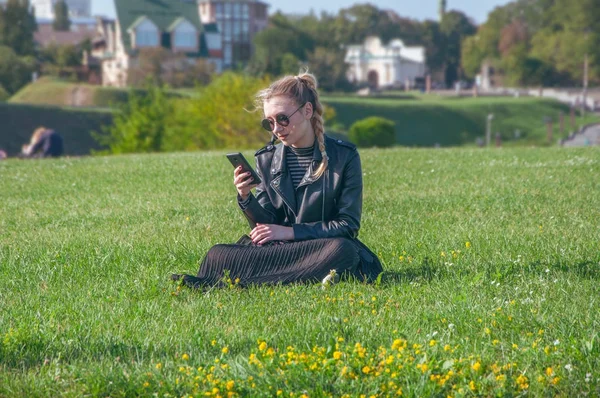 Hermosa chica rubia se sienta en un césped verde y mira en un teléfono inteligente — Foto de Stock