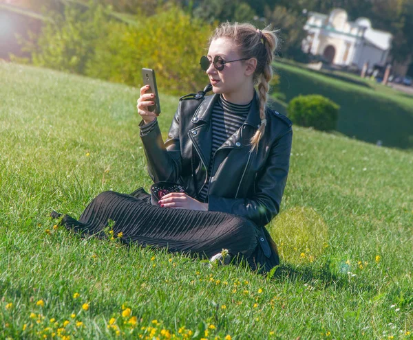 Hermosa chica rubia se sienta en un césped verde y mira en un teléfono inteligente — Foto de Stock