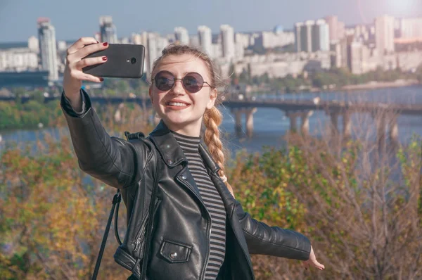 Hermosa chica rubia se para en el fondo de la ciudad y hace selfie — Foto de Stock