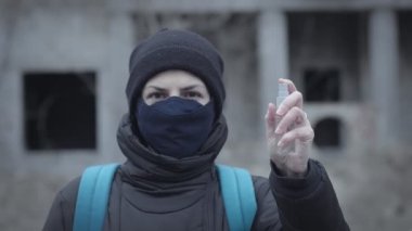 Koruyucu maskeli genç bir kadın, terk edilmiş bir binanın arka planına antiseptik sprey sıkar. Pandemik koruma, hava kirliliği