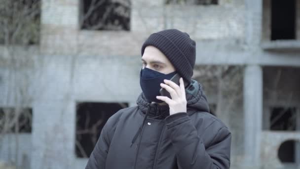 Een man met een beschermend masker spreekt telefonisch tegen de achtergrond van een verlaten gebouw in een industrieel postapocalyptisch landschap. Pandemische bescherming — Stockvideo