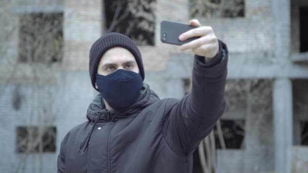 Een jonge man met een beschermend masker schreeuwt video met zijn smartphone tegen de achtergrond van een verlaten gebouw in een industrieel post-apocalyptisch landschap — Stockvideo