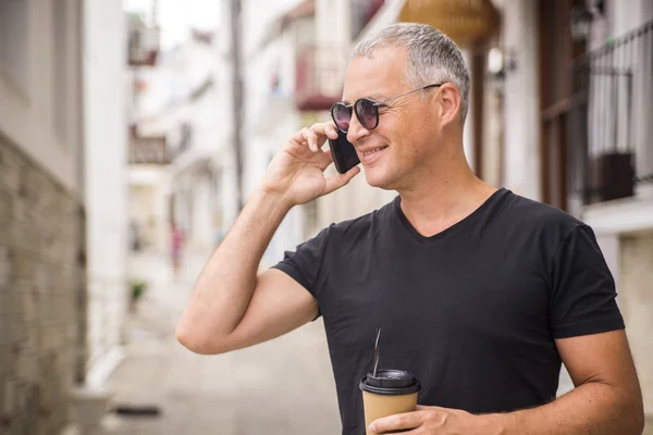 Stadsleven. Knappe jongeman in slimme vrijetijdskleding wandelen langs de straat terwijl koffiekopje en slimme telefoon — Stockfoto