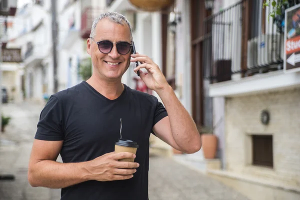 Talking with friend. Handsome man in smart casual wear carrying coffee cup and talking on mobile phone while walking along the street