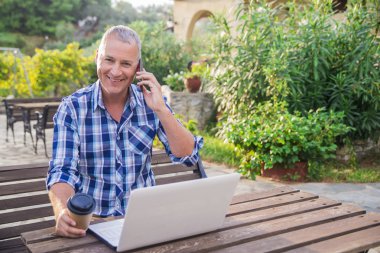 Ofis kaçtı. Çalışma dizüstü içme kahve güneş ışığı ve zemin üzerine yeşil Teras etrafında elektronik becerikli alet ile naturalcountry tarzı ahşap masa başında oturan adam iş tarzı giyinmiş