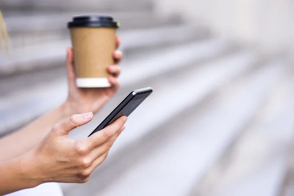 Mulher segurar xícara de café e telefone inteligente na rua, com espaço para texto . — Fotografia de Stock