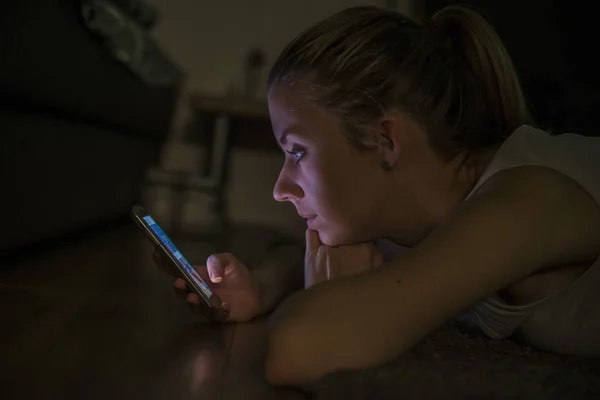 Портрет молодої жінки, що лежить на килимі з мобільним телефоном вдома вночі — стокове фото