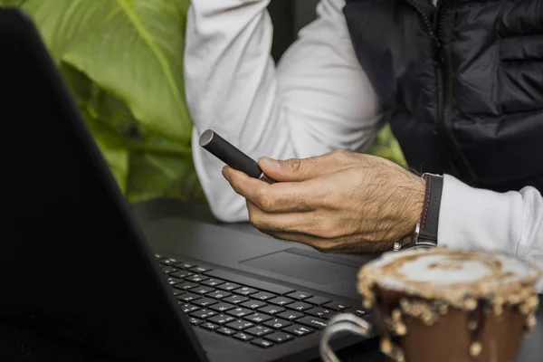 Cropped image of modern man typing something on laptop, with watch and e-cigarette. man with laptop sitting in a cafe outdoor and smoking electronic cigarette. Handsome man working in cafe.