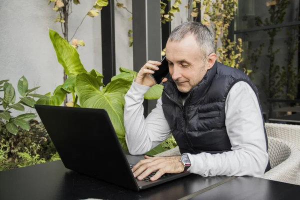 Joven hombre de negocios bebiendo una taza de café mientras está sentado en una mesa de la terraza de una cafetería usando una computadora portátil y haciendo una llamada telefónica en su teléfono inteligente, al aire libre. Hablando con amigos. Joven confiado hablando por teléfono móvil y sonriendo — Foto de Stock