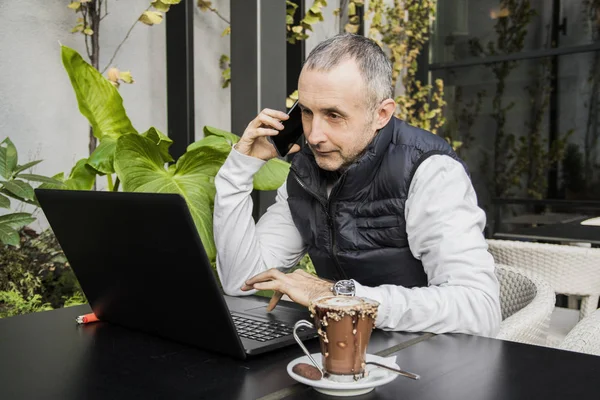 一杯のコーヒーを飲みながらコーヒー ショップに座って青年実業家テラス テーブル ラップトップ コンピューターを使用し、スマート フォン、屋外の電話をかけます。友人と話しています。自信を持って若い男が携帯電話で話していると笑顔 — ストック写真