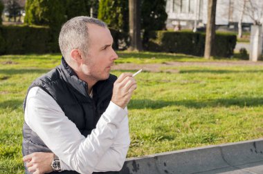 Bir ağacına bir parkta otururken, bir sigara ile rahatlatıcı genç iş adamı. İş adamı iyi giyimlisin sokak kaldırımda otururken sigara, madde bağımlılığı, bağımlılık, insanlar ve kötü alışkanlıkları kavramı - kadar yakın genç adam Sigara İçilmeyen açık