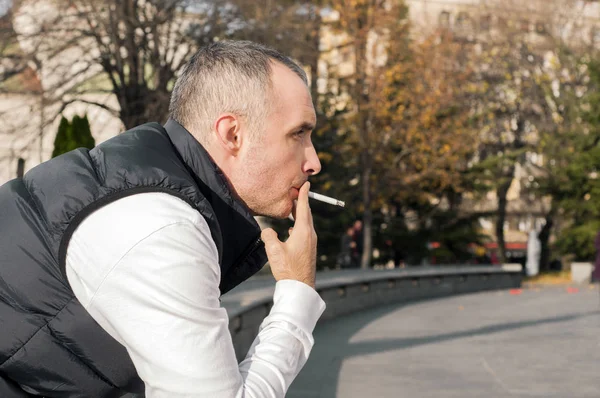 Schöner, stylischer junger Mann, der draußen in urbaner Umgebung raucht und wegschaut. schöner weißer Mann, der an einem sonnigen Tag draußen eine Sigarette raucht — Stockfoto