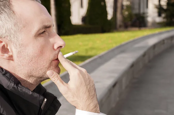 Joven hombre de negocios sentado contra un árbol en un parque, relajándose con un cigarrillo. Hombre de negocios bien vestido fumando sentado en una acera de la calle, abuso de sustancias, adicción, personas y malos hábitos concepto - primer plano de hombre joven fumando cigarrillo al aire libre — Foto de Stock