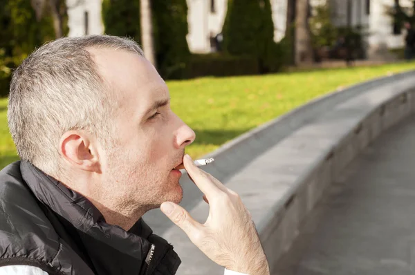 Schöner, stylischer junger Mann, der draußen in urbaner Umgebung raucht und wegschaut. schöner weißer Mann, der an einem sonnigen Tag draußen eine Sigarette raucht — Stockfoto