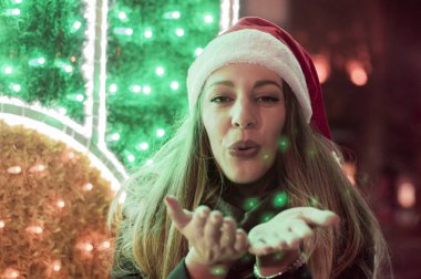 Noel, x-mas, insanlar, mutluluk kavramı - avucunda üfleme kış giysileri içinde mutlu bir kadın. Mutlu kız kış Aydınlatmaları Noel ağaçları ve parlak carousel ile arasında bir öpücük kış kap içinde gönderir.