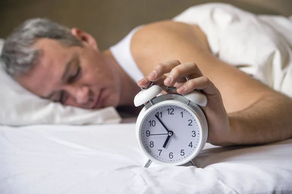 Man lying in bed turning off an alarm clock in the morning at 7am. Attractive man sleeping in his bedroom. Annoyed man being awakened by an alarm clock in his bedroom — Stock Photo, Image
