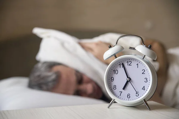 Hombre Despertado acostado en la cama apagando un despertador en la mañana a las 7 am. Hombre tratando de dormir, cuando suena el despertador —  Fotos de Stock