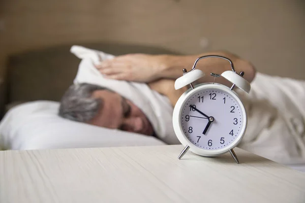 O homem acordado deitado na cama a desligar um despertador de manhã às 7h. Homem a tentar dormir, quando o despertador toca — Fotografia de Stock