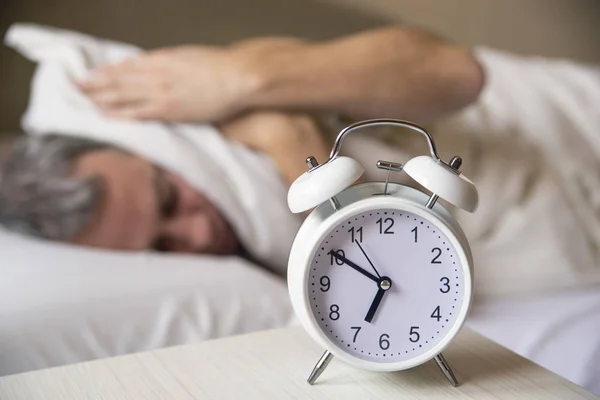 Slapende man verstoord door wekker vroeg in de ochtend. Sleepy jongeman bedekking oren met kussen als hij kijkt naar de wekker in worden — Stockfoto