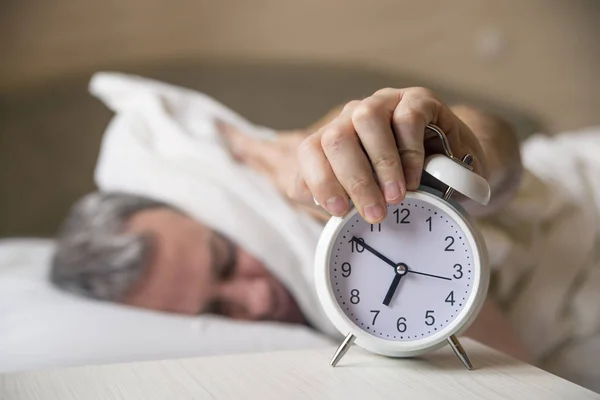 Sover man störd av väckarklockan tidigt på morgonen. Sömnig ung man som täcker öronen med kudde vara som han ser på väckarklocka i — Stockfoto