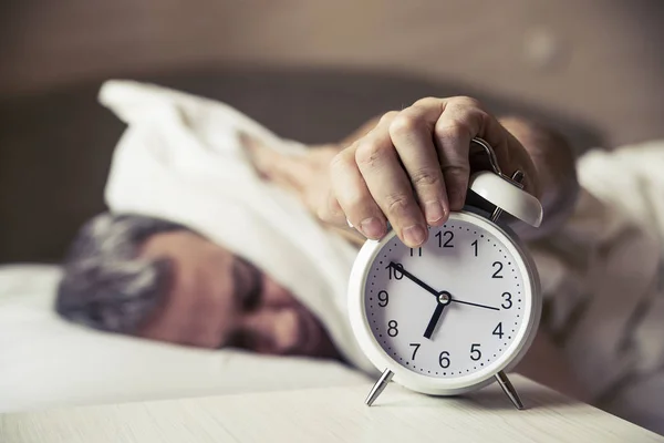 Sleepy jongeman die oren met kussen als hij naar de wekker in bed kijkt. slapende man verstoord door wekker vroeg in de ochtend. Gefrustreerde man luisteren naar zijn wekker terwijl u ontspant op zijn bed — Stockfoto