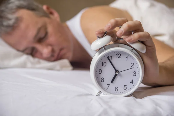 Älterer Mann schlafend im Bett mit Wecker im Vordergrund im Schlafzimmer. Erschöpfter Mann wird von einem Wecker in seinem Schlafzimmer geweckt. aufgewacht lizenzfreie Stockfotos