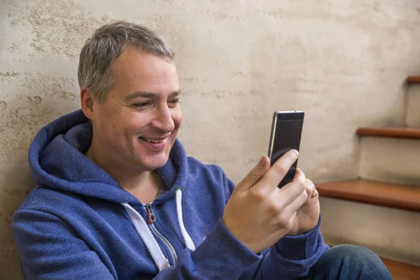Fyrtio år gammal affärsman sittplatser på trappa inuti kontorsbyggnaden tittar på en mobiltelefon. glad affärsman leende medan du läser hans smartphone. — Stockfoto