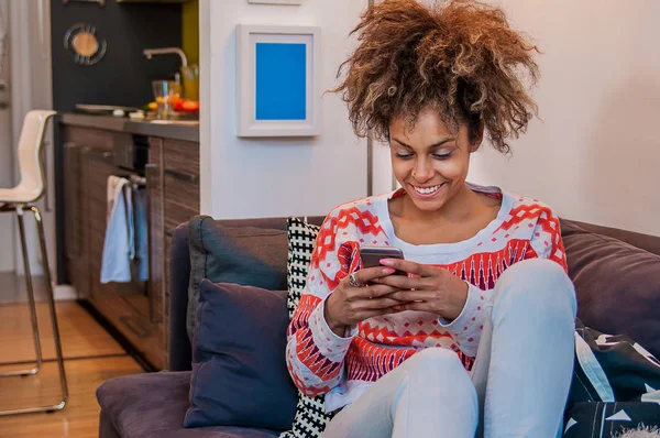 Афроамериканка использует смартфон дома. Черные. Женщина пишет смс с мобильного телефона, сидя на диване — стоковое фото