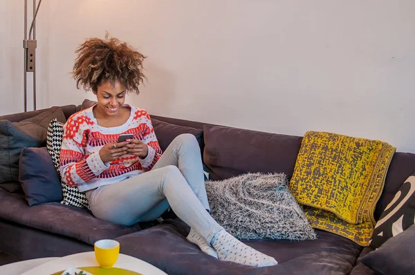 Afrikanisch-amerikanische Frau mit Smartphone zu Hause. Schwarze Menschen. Frau schreibt SMS mit Handy, während sie auf der Couch sitzt — Stockfoto