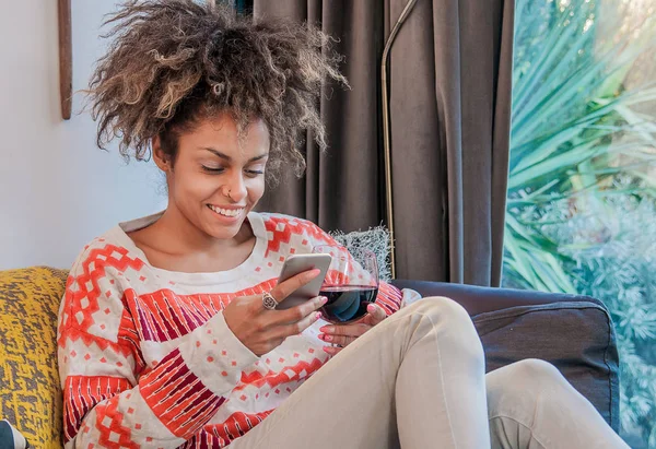 Lächelnde afrikanisch-amerikanische Frau, die auf dem Sofa sitzt und SMS liest. lächelnde junge Frau zu Hause, die es sich auf der Couch gemütlich macht, sie benutzt Smartphone und SMS, Technologie und Kommunikationskonzept — Stockfoto