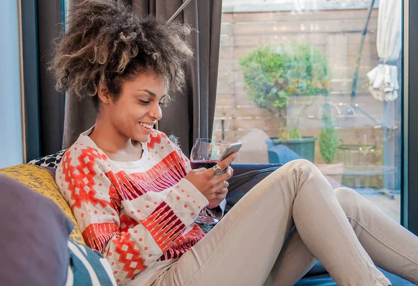 微笑的非洲裔美国人的女士坐在沙发上，阅读文本消息。微笑在家里在沙发上的年轻女子，她使用智能手机和短信、 技术和通信的概念 — 图库照片