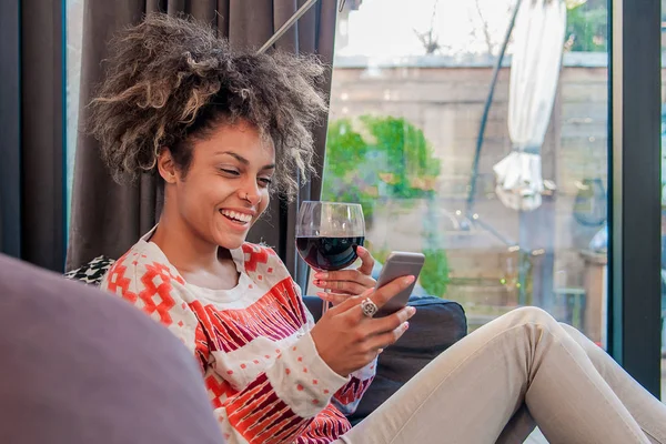 Красивая женщина в своей гостиной лежит на диване и отправляет сообщения. Портрет африканской женщины, смотрящей на мобильный телефон и наслаждающейся бокалом вина — стоковое фото