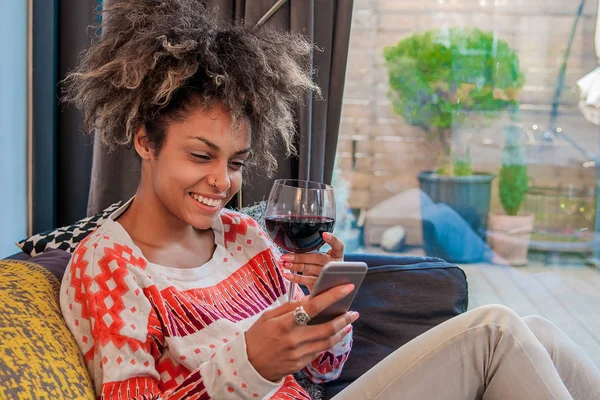 漂亮的女人，在她的客厅里，躺在沙发上发送消息。看着手机，享受杯酒非洲女人的画像 — 图库照片