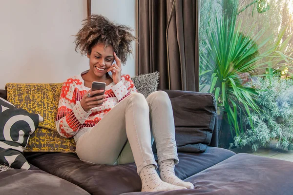 SMS an ihn. attraktive junge Frau mit Smartphone und Lächeln. Porträt einer attraktiven jungen Frau, die SMS auf dem Handy liest, während sie es sich zu Hause auf dem Sofa gemütlich macht — Stockfoto