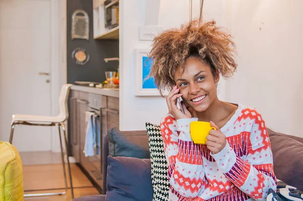 Bonjour sans soucis. Belle jeune femme métissée tenant une tasse de café et parlant sur le téléphone portable avec le sourire tout en étant assis sur le coach — Photo