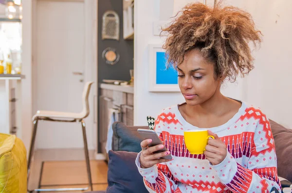 Portrait d'une jeune femme noire souriante assise sur un canapé à la maison avec un téléphone portable. Belle femme afro-américaine avec une coiffure afro crépus textos sur son mobile — Photo