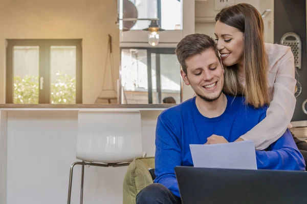 Foto de alegre pareja joven y cariñosa utilizando el ordenador portátil y el análisis de sus finanzas con documentos. Mira los papeles.. — Foto de Stock