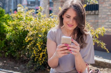 Bir bankta oturan ve cep telefonuyla genç kadın. Attractivk yeşil bir odaklanmamış arka plan ile. Neşeli kadın manifatura bir parkta onun smartphone ile