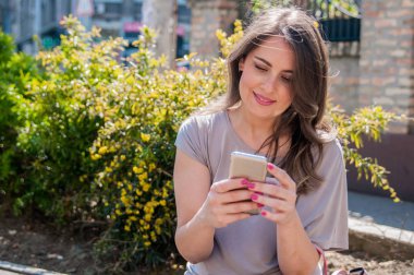 Bir bankta oturan ve cep telefonuyla genç kadın. Attractivk yeşil bir odaklanmamış arka plan ile. Neşeli kadın manifatura bir parkta onun smartphone ile