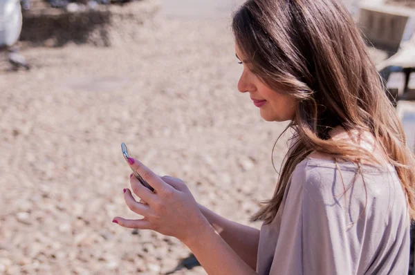 Mujer bonita usando su teléfono móvil mientras está sentada en un banco de madera. El estilo casual - los vaqueros y la camisa. Mujer joven feliz usando el teléfono inteligente en un día soleado — Foto de Stock