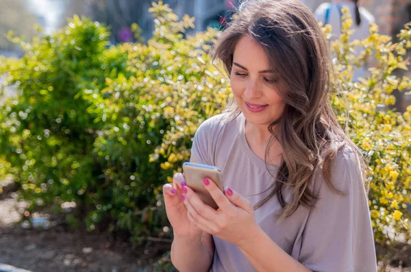 Un retrato de una hermosa mujer sonriente enviando mensajes con su teléfono. Mujer atractiva usando el teléfono celular al aire libre . — Foto de Stock